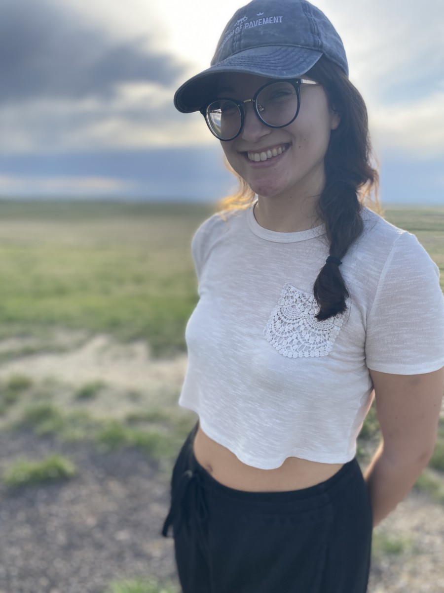 Amy suto smiling while standing in a grass field with some blue sky and clouds behind her as she wears a cropped white shirt, glasses, a baseball hat and her hair in a loose braid