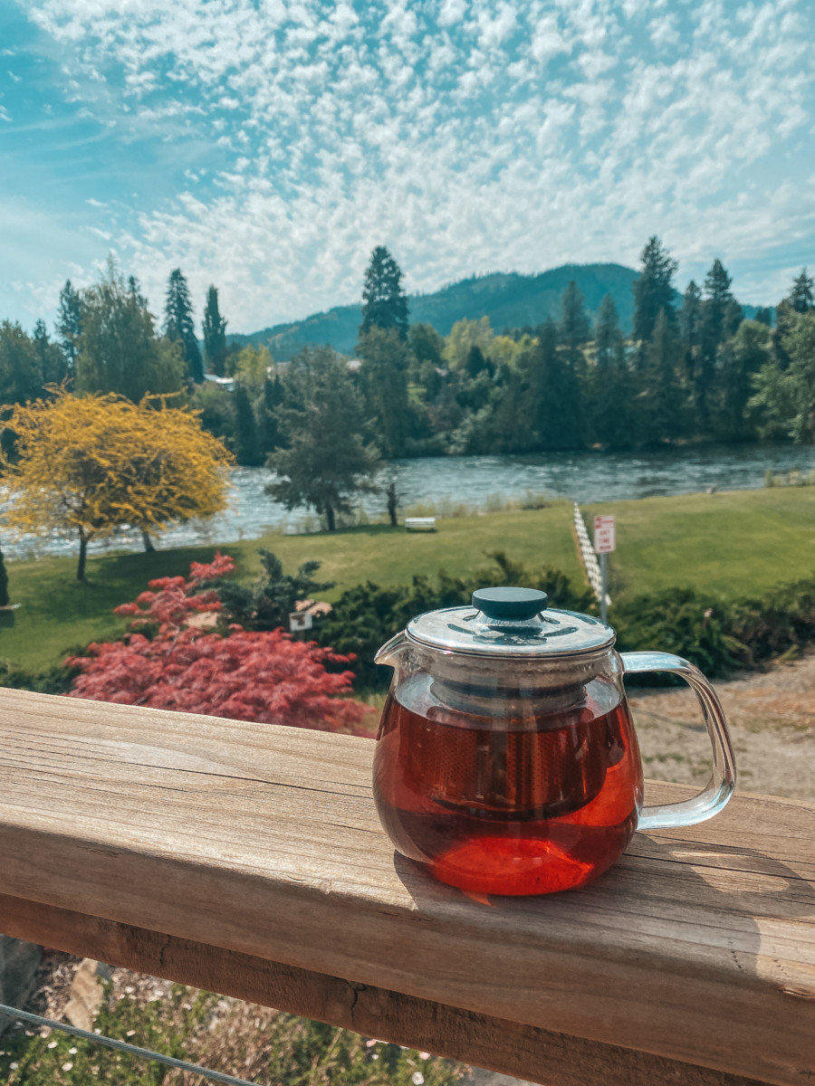 a teapot full of red tea sits on the railing of a patio that overlooks a yard full of trees and a lake
