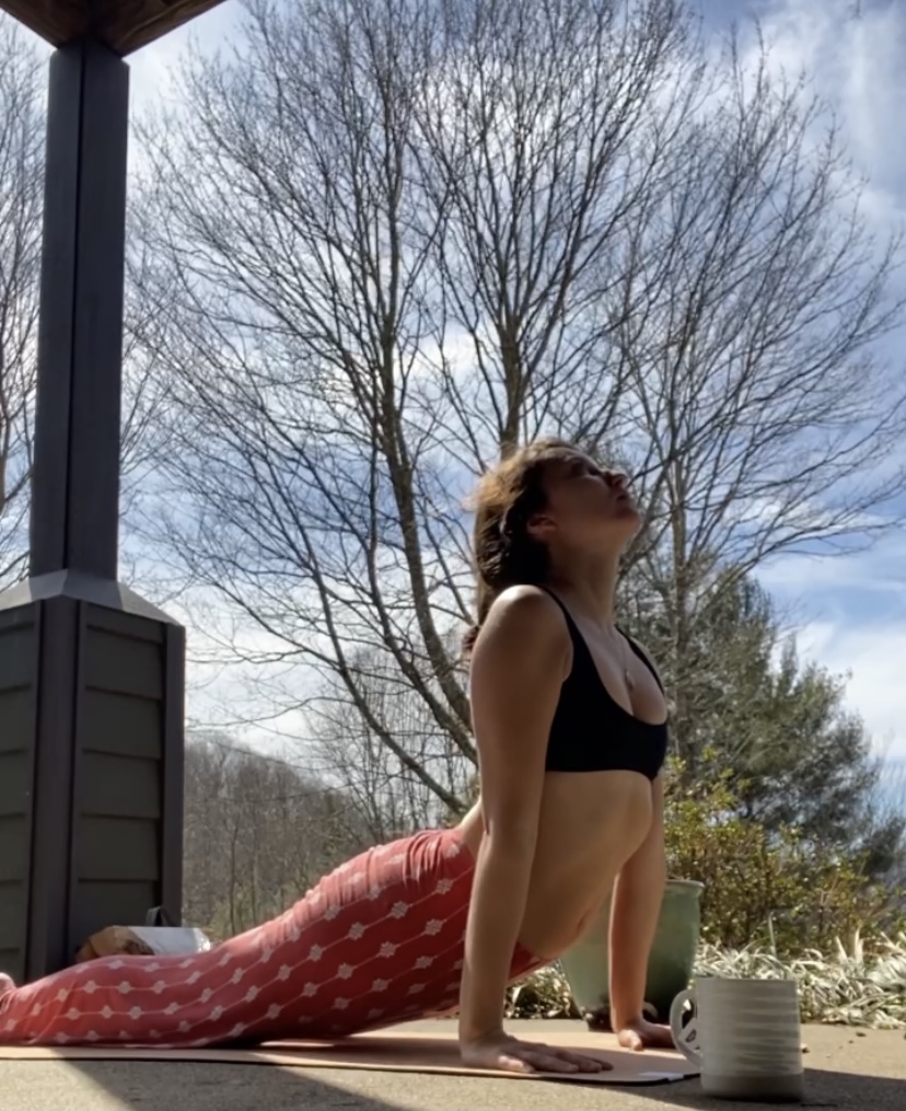 Amy Suto performs a yoga move on a deck on a sunny day with trees behind her