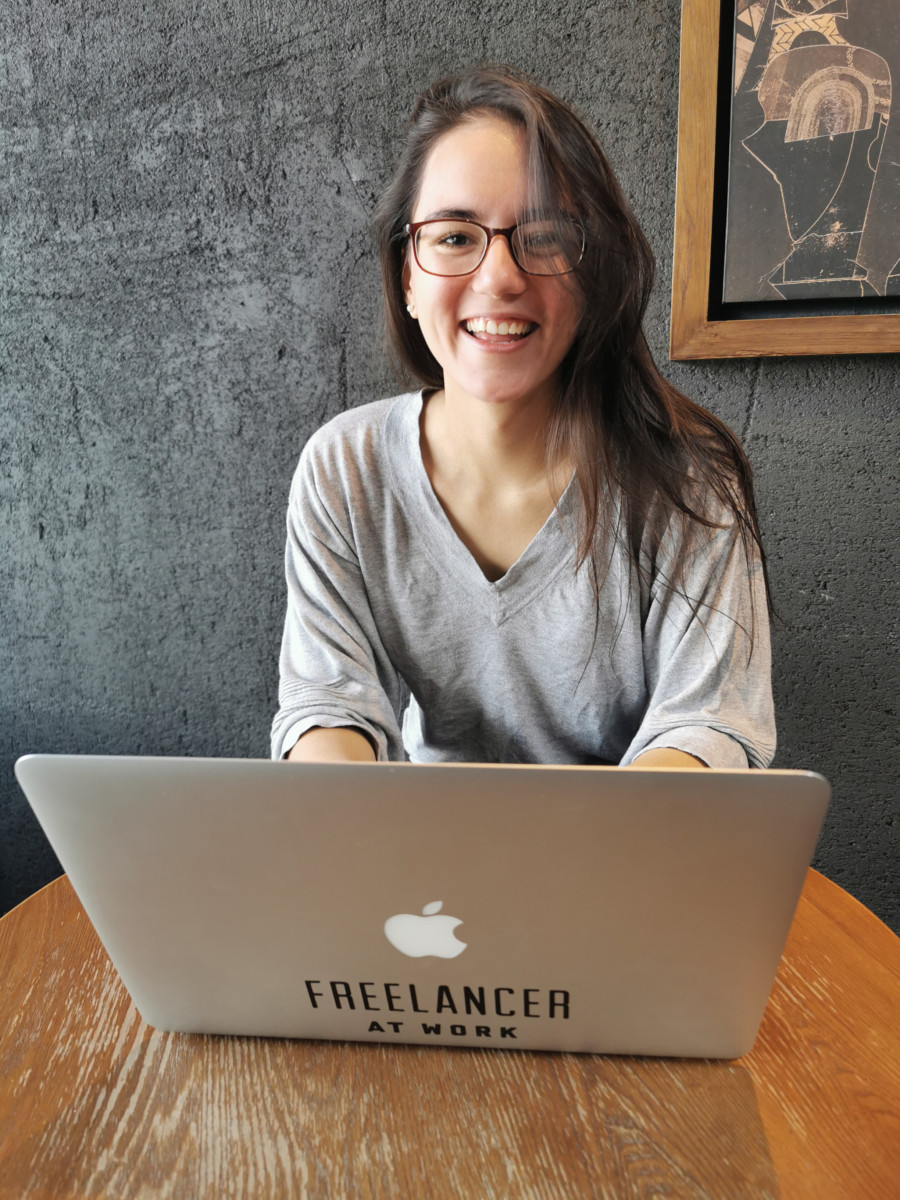 Deya sits at a table with her laptop open in front of her smiling and wearing glasses