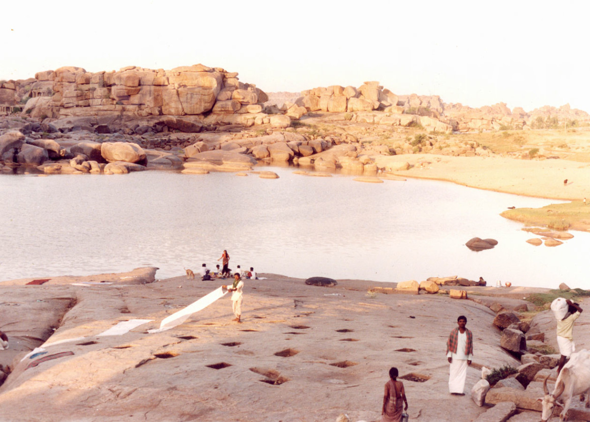 large boulder formations in hampi, india while people stand on the rocks that slop down to a lake