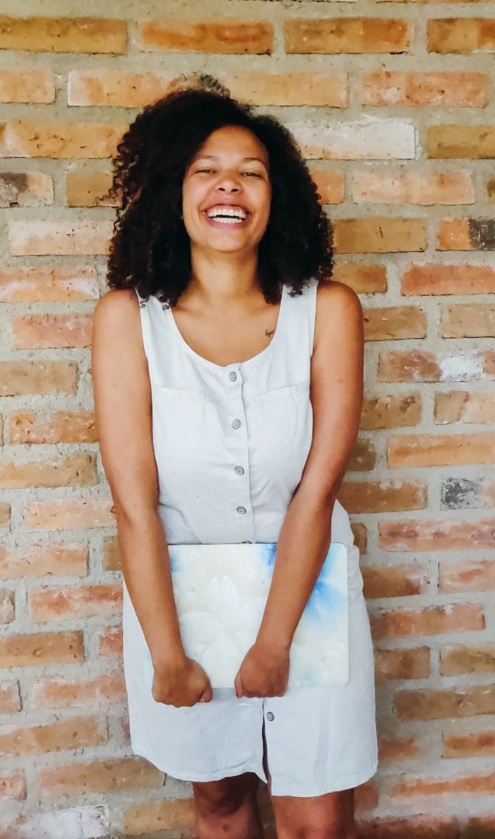 Felly day stands in front of a brick wall while smiling and holding her laptop