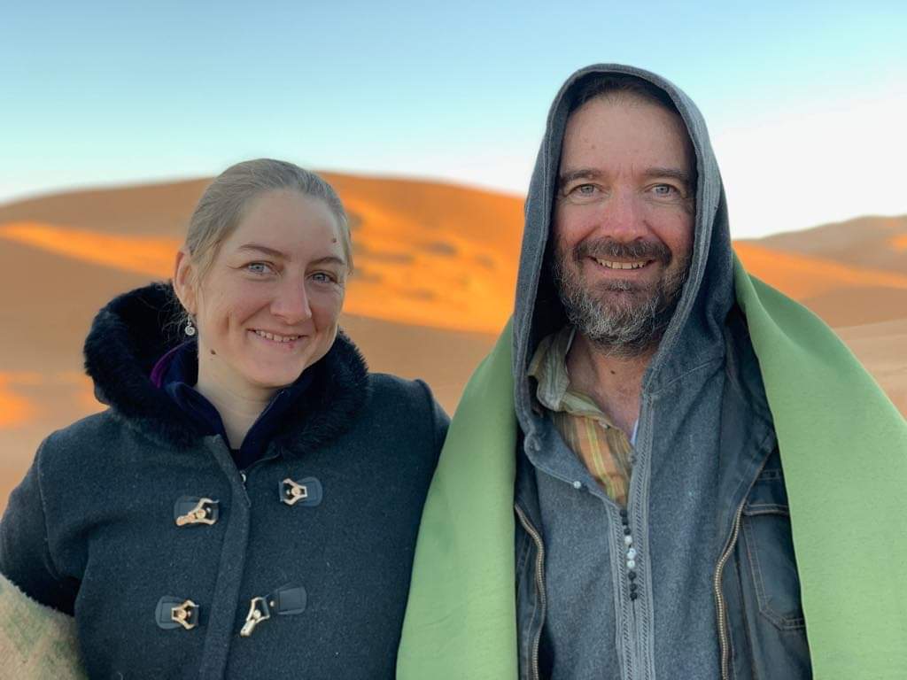 Tim and Kassandra Marsh wearing heavy coats in the Sahara desert of Morocco with the sun shining over a sand dune behind them