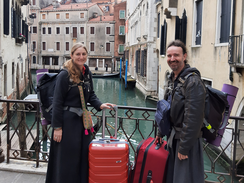 Tim and Kassandra Marsh stand on a bridge over a canal in Venice, each holding one large suitcase and one backpack