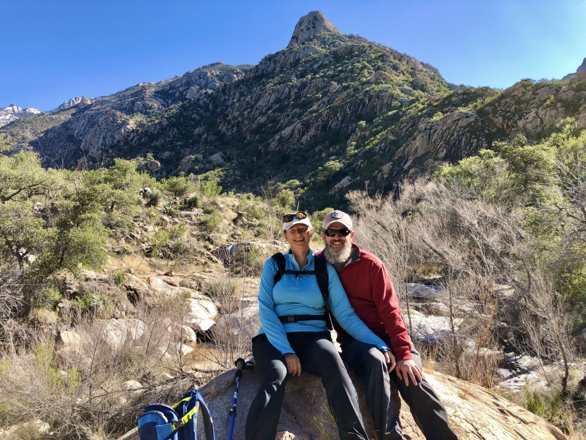 in Tuscon, AZ, Julie and Sean chickery sit together on a rock with their backpack on the ground next to them and a large mountain in the background