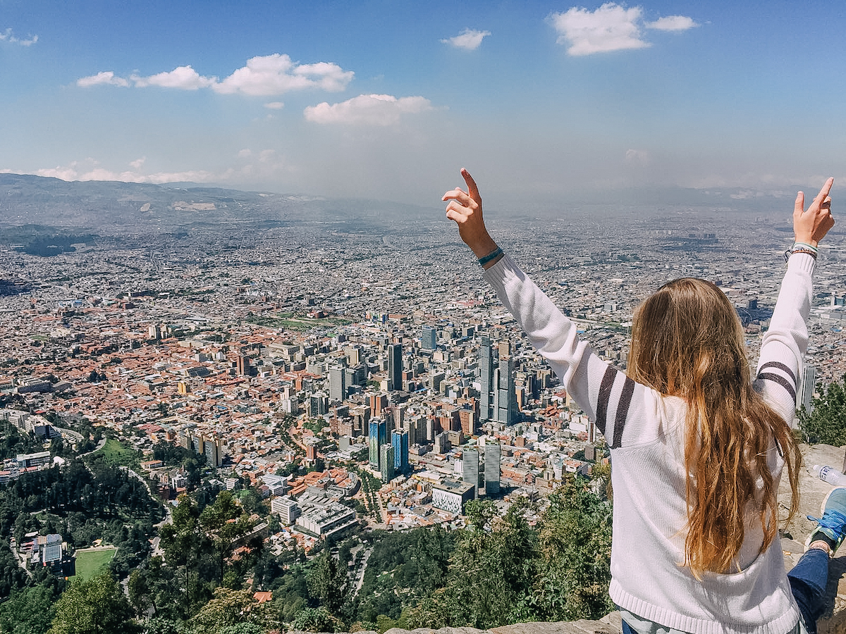 Anny Woolridge with her back to the camera and her arms in the air while standing atop a tall hill that overlooks Bogota, Columbia