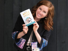 Meredith Noble stands holding a book she wrote in her hands near her face while smiling