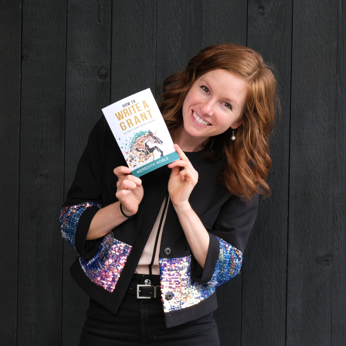 Meredith Noble stands holding a book she wrote in her hands near her face while smiling