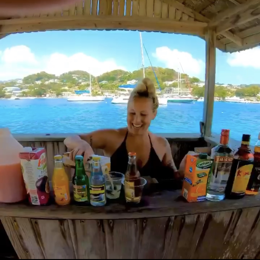 Angela Faith pouring a drink while standing behind a tiki bar full of bottles with the ocean behind her