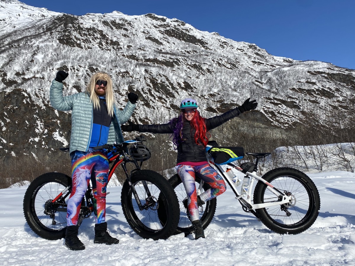 meredith noble and her boyfriend lucas both wear unicorn pants standing in front of their large bicycles in the snow while a large, snowy mountain sits behind them