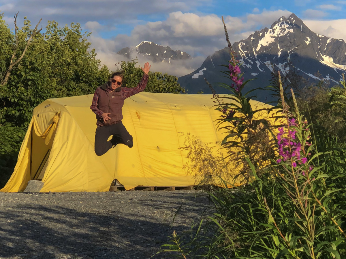 Meredith Noble jumps in the air in front of a huge yellow tent in Alaska with fireweek in the foreground and a snow-capped mountain behind the yellow tent