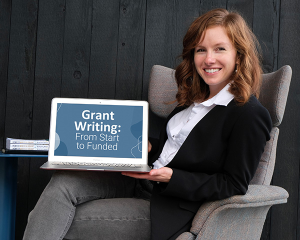 Meredith noble sits facing to the right in a large chair with her legs crosses holding a laptop with a screen saying "grant writing: from start to funded."