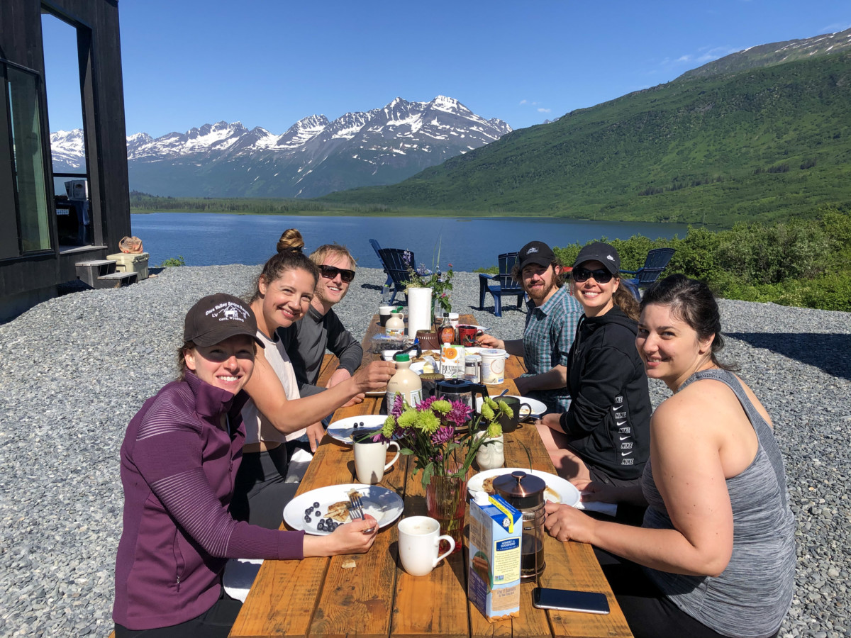 Meredith noble sits at a long picnic table with five other people eating a large spread of breakfast food while sitting next to a lake in alaska