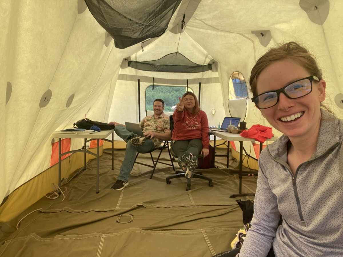 Meredith noble sits in the foreground with two other cowers in the back sitting at desks in a large white tent where they work in alaska