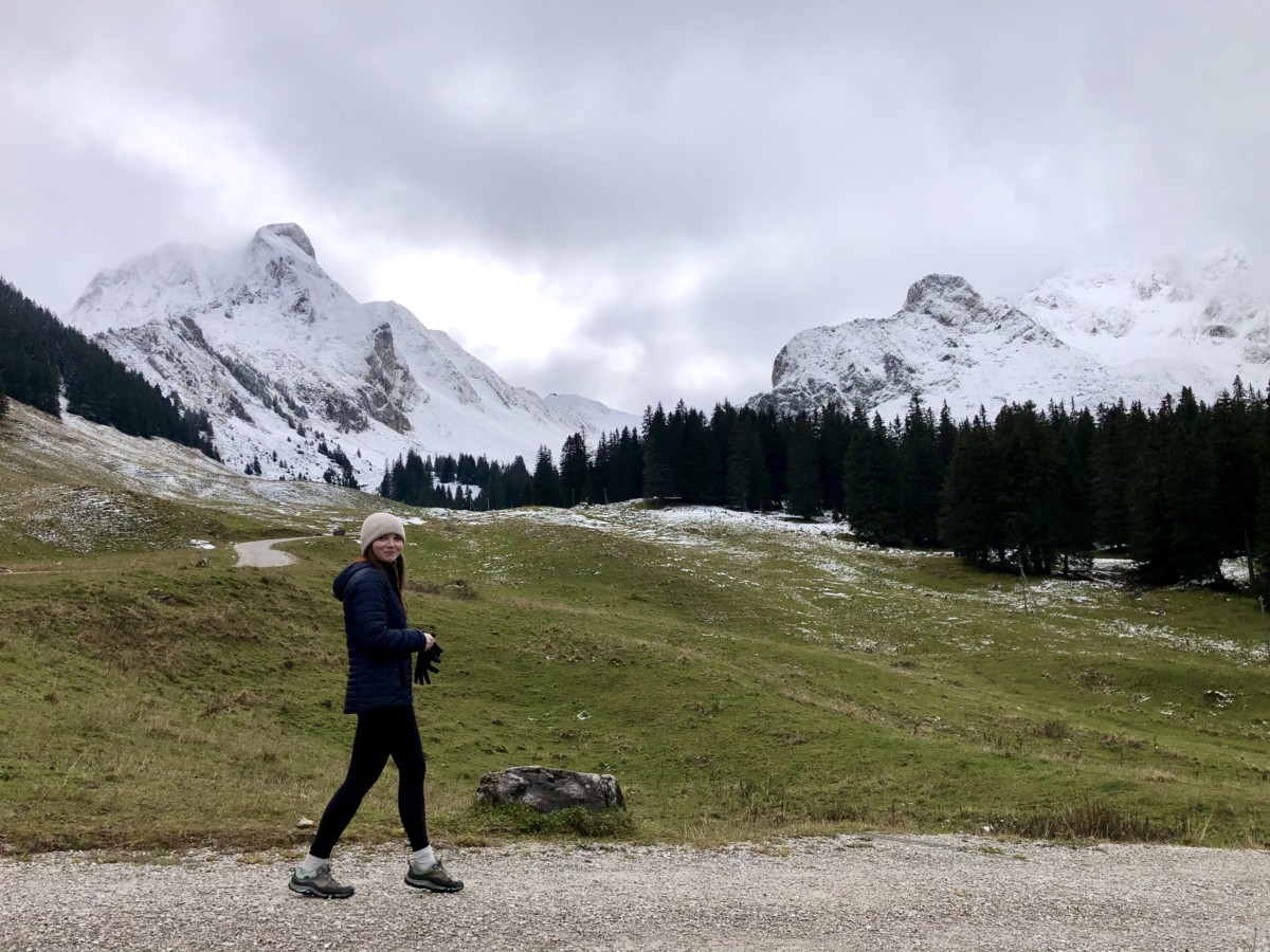 Sarah archer walks on a path in front of snow-capped mountains near a field of green grass