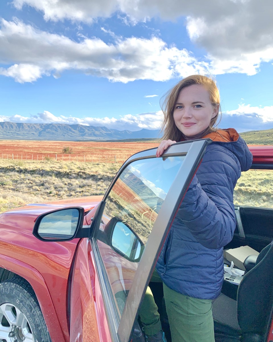 Sarah Archer stands between a red car and the driver's door wearing a puffy jacket in Patagonia with a field and blue sky behind her