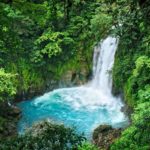 A rushing waterfall flows into an emerald blue pool below with lush vegetation and cliffs around it