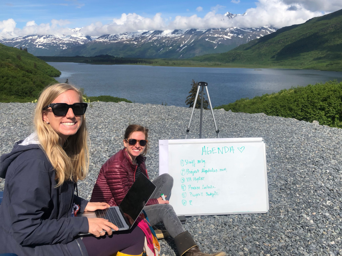 Meredith Noble sits in the gravel ground with co-worker Alex who sits in a chair with a laptop on her lap while both are located next to mere lake in Alaska