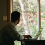 Man in gray hoodie using laptop while looking to the outside