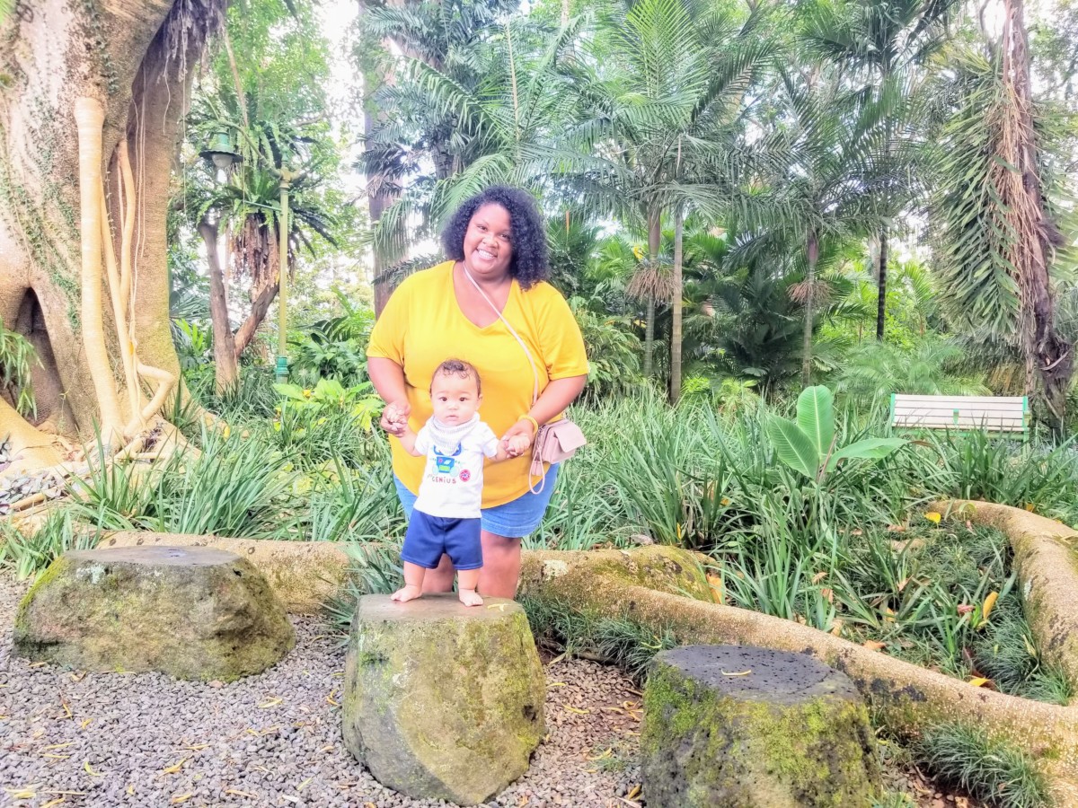 A small boy stands on a rock in a forest while a woman stands behind him holding his shoulders