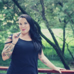 A women stands with one arm on a railing and another holding a vape pen in front of a green landscape with trees