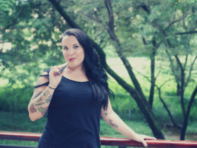 A women stands with one arm on a railing and another holding a vape pen in front of a green landscape with trees