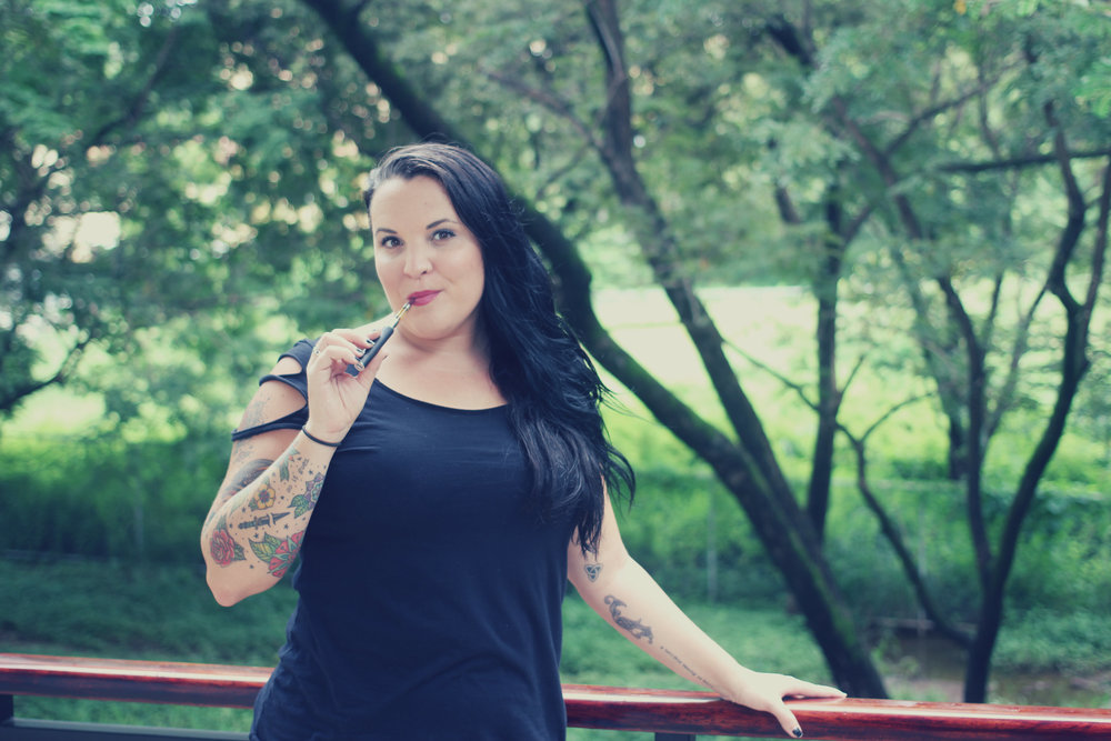 A women stands with one arm on a railing and another holding a vape pen in front of a green landscape with trees