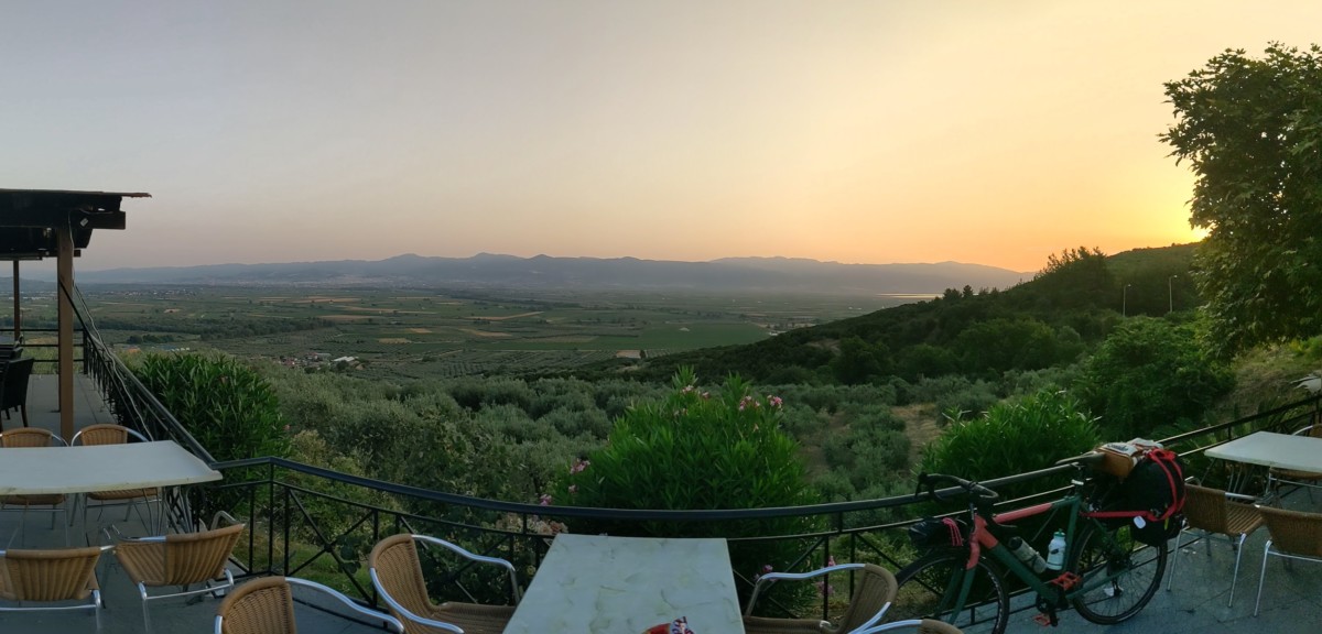 empty tables and chairs sit on a patio along with a bicycle leaning against a railing of the dining area that overlooks a vast area with farm land and plants