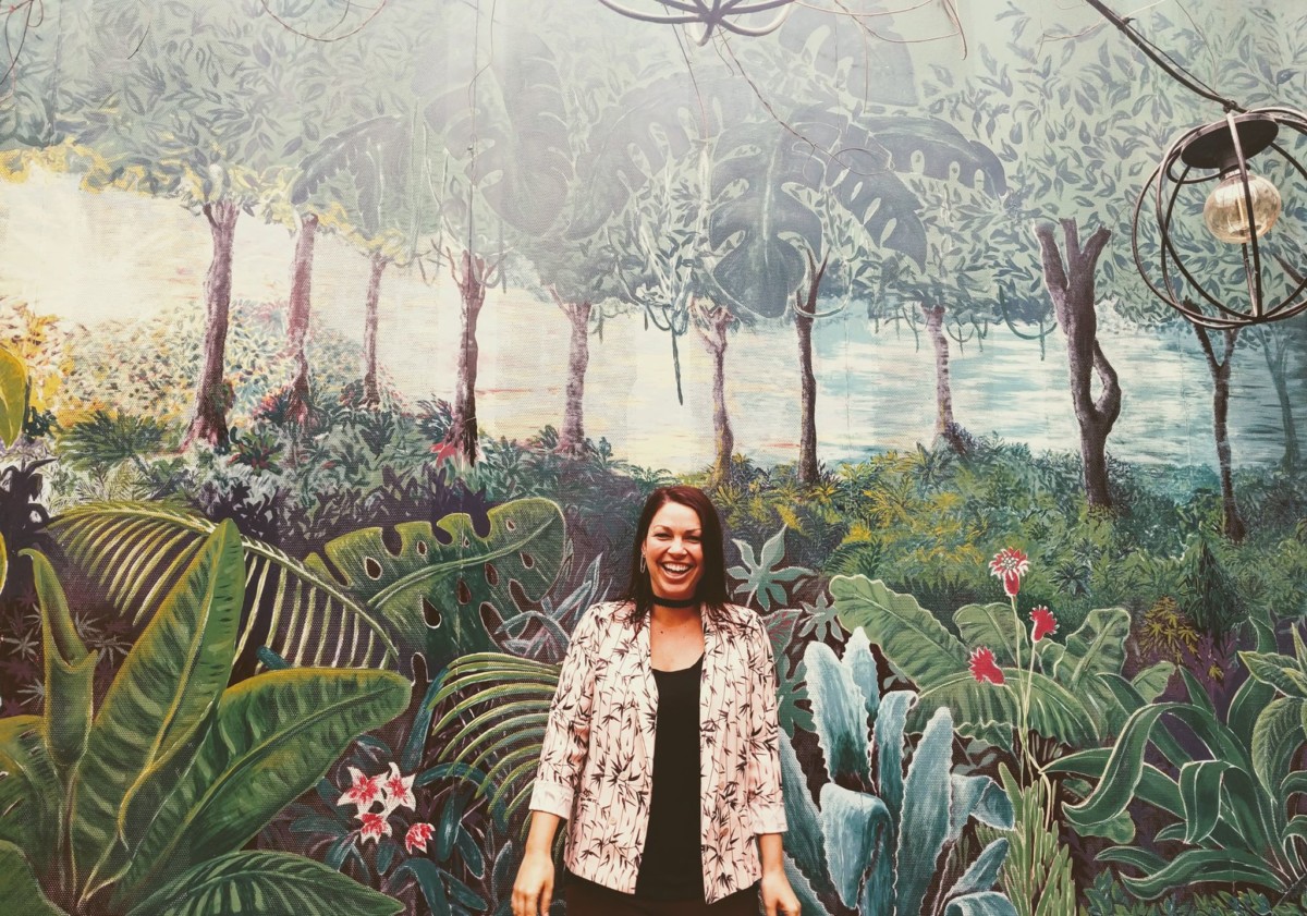 Lolly stands smiling in front of a wall mural painted with tropical vegetation