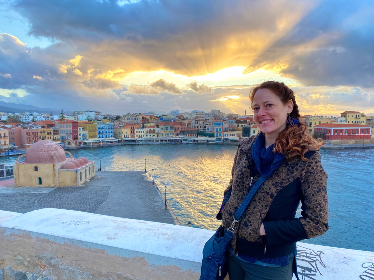 Nora Dunn stands in front of the setting sun in Crete with many buildings and the ocean behind her