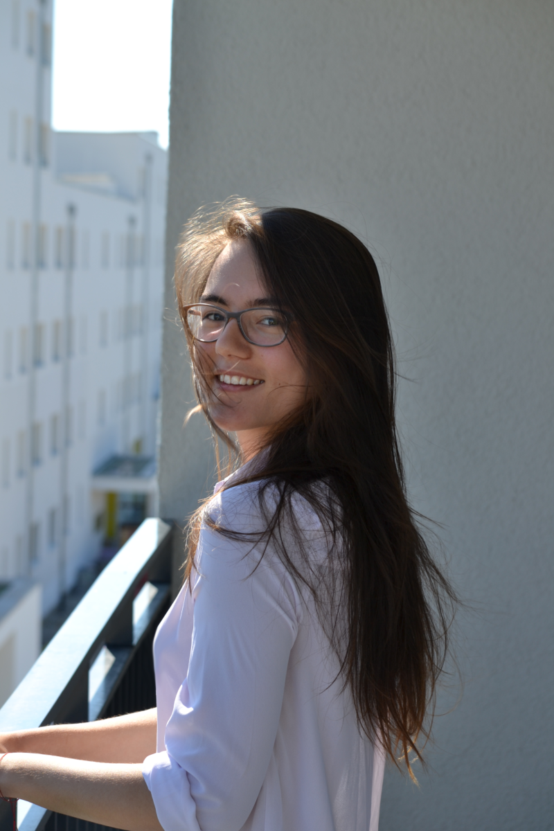 Deya stands on a sunny balcony holding the railing while turned slightly sideways smiling