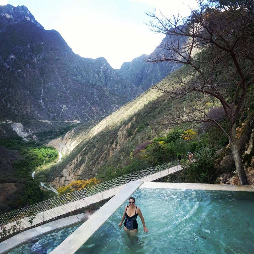 Lolly stands in a swimming pool that is next to a large and long suspension bridge with tall mountains surrounding the area