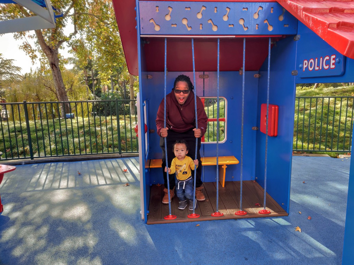A small boy stands in front of a woman on a playground in a fake police cell