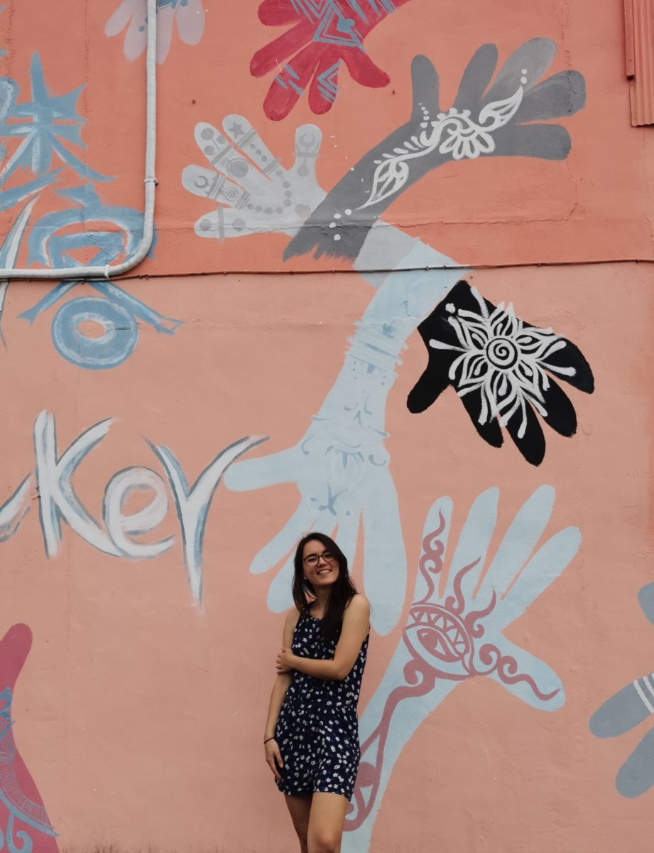 Deya stands with one arm across her body to hold her other arm while standing in front of a coral colored wall with hands painted on it