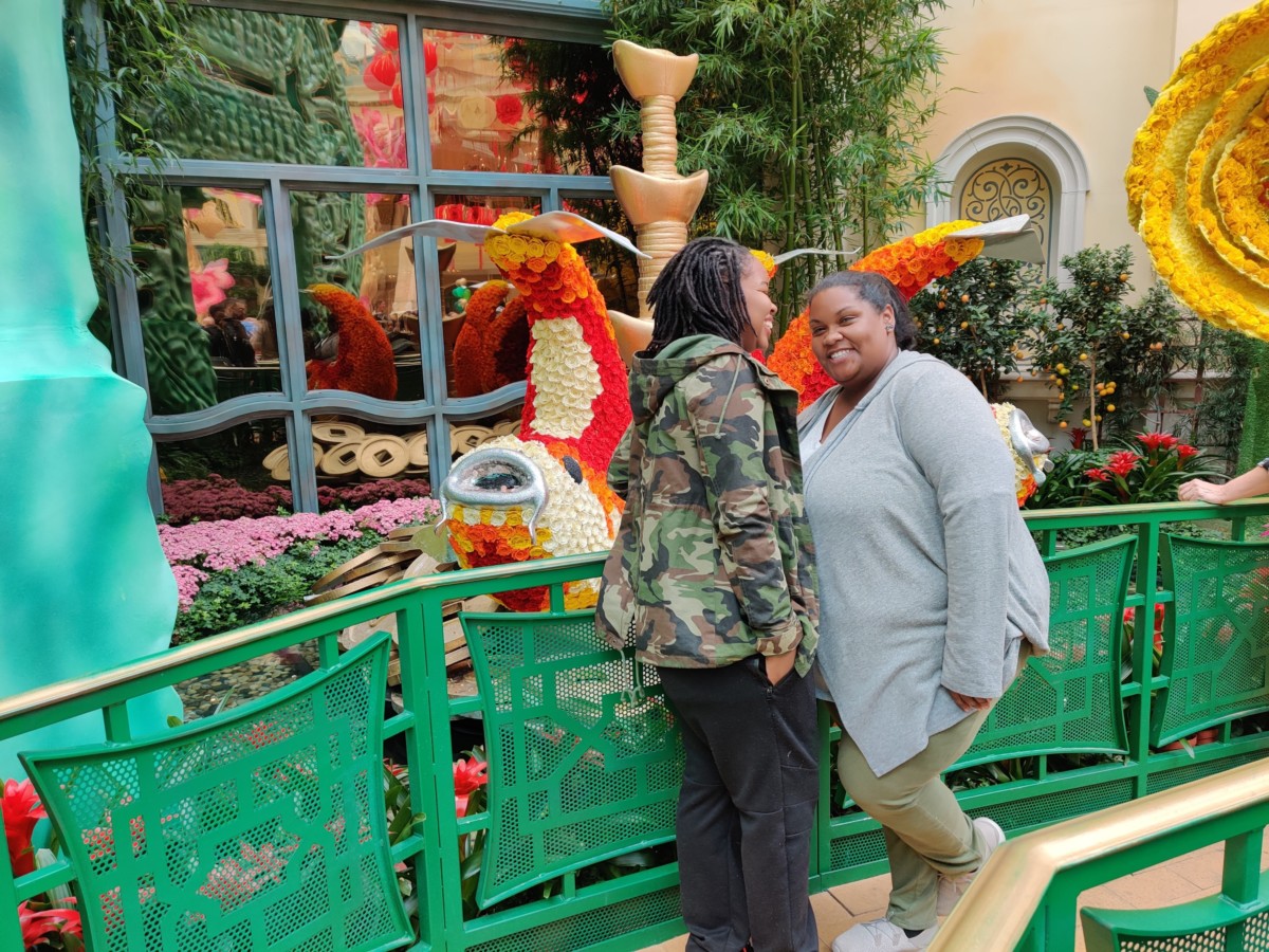 Two women stand facing each other smiling with ornate flower decorations in the shape of birds behind them