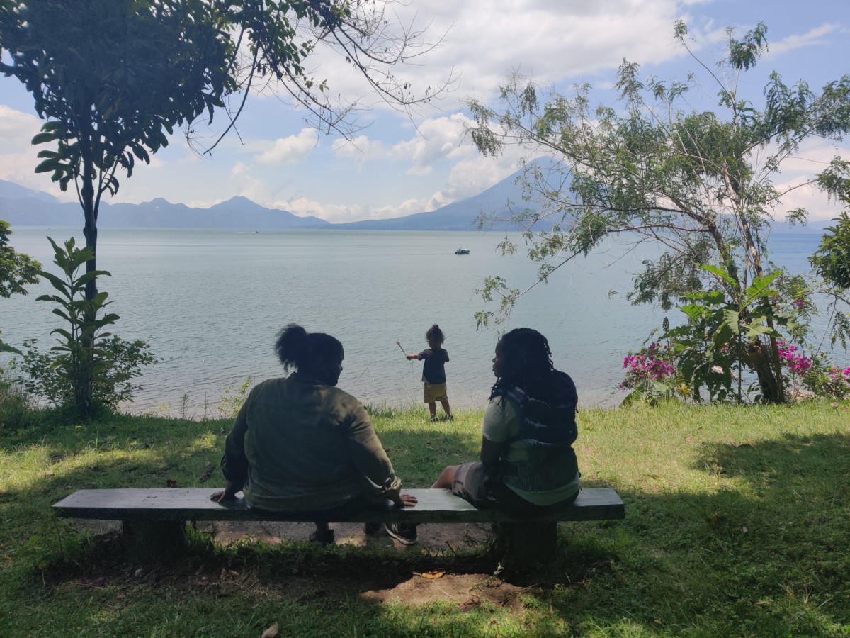 two women sit on a bench facing a lake while their small son plays near the lake's shore