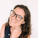 nina clapperton with a white background while she poses with her left hand on her neck, smiling and wearing glasses