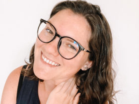 nina clapperton with a white background while she poses with her left hand on her neck, smiling and wearing glasses