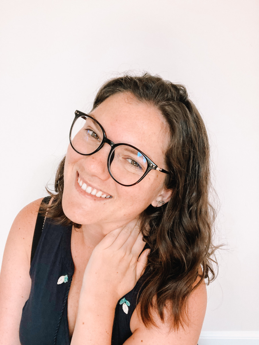 nina clapperton with a white background while she poses with her left hand on her neck, smiling and wearing glasses