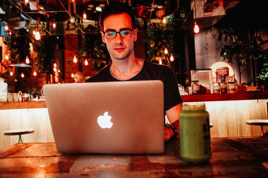 Blake Miner sits in a cafe working with his laptop in front of him and a green drink next to his computer while sitting in front of a bar with coffee implements and plants hanging from the celing