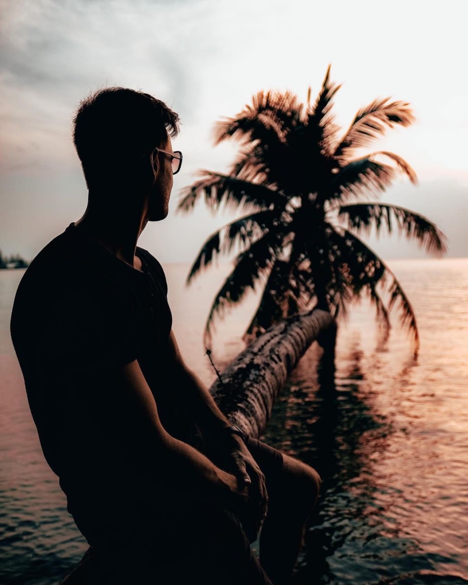 Blake Miner sits on the trunk of a palm tree at sunset that has grown horizontally over the ocean