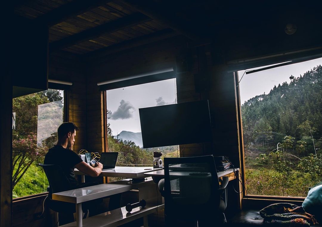 Blake Miner sits in a room with many large windows that look into nature while he works on a laptop at a desk