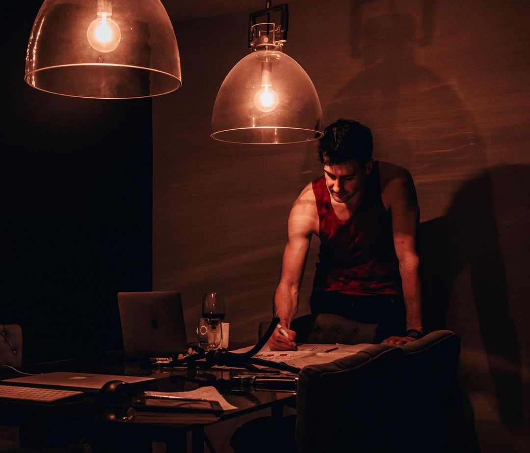 Blake Miner stands at a table with paperwork strewn across it while two lights hang above illuminating the otherwise dark room