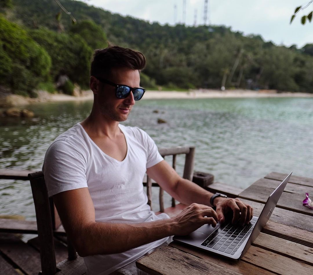 Blake Miner sits at a wooden table working on his laptop next to a tropical beach with a sandy shore and tropical vegetation