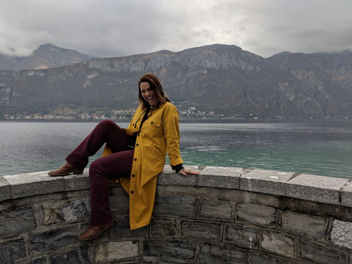 Lolly sits with one leg up on a stone wall in front of lake como on a grey day while wearing a long yellow rain jacket