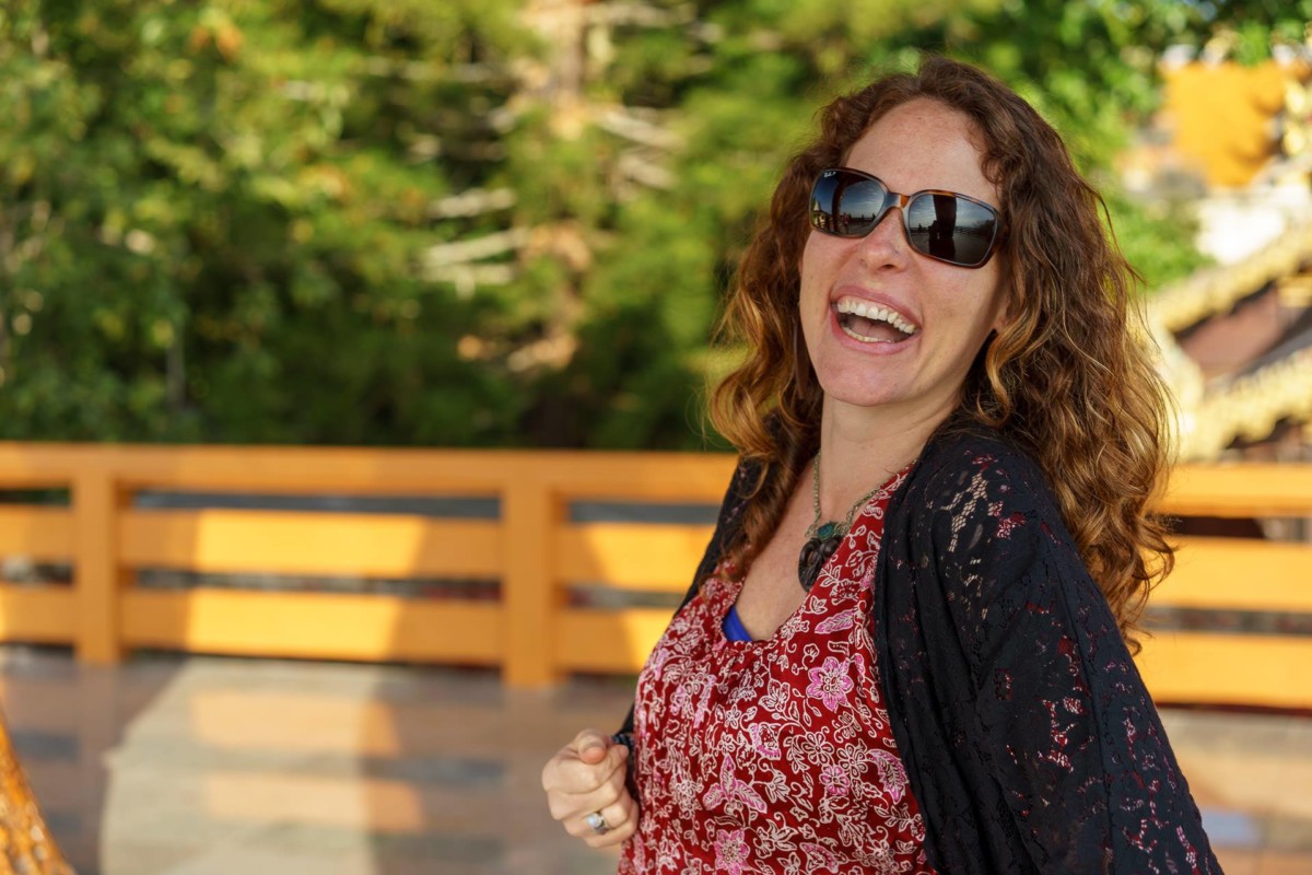 A female wearing sunglasses smiles and laughs while outside in front of a yellow fence with green plants behind the fence