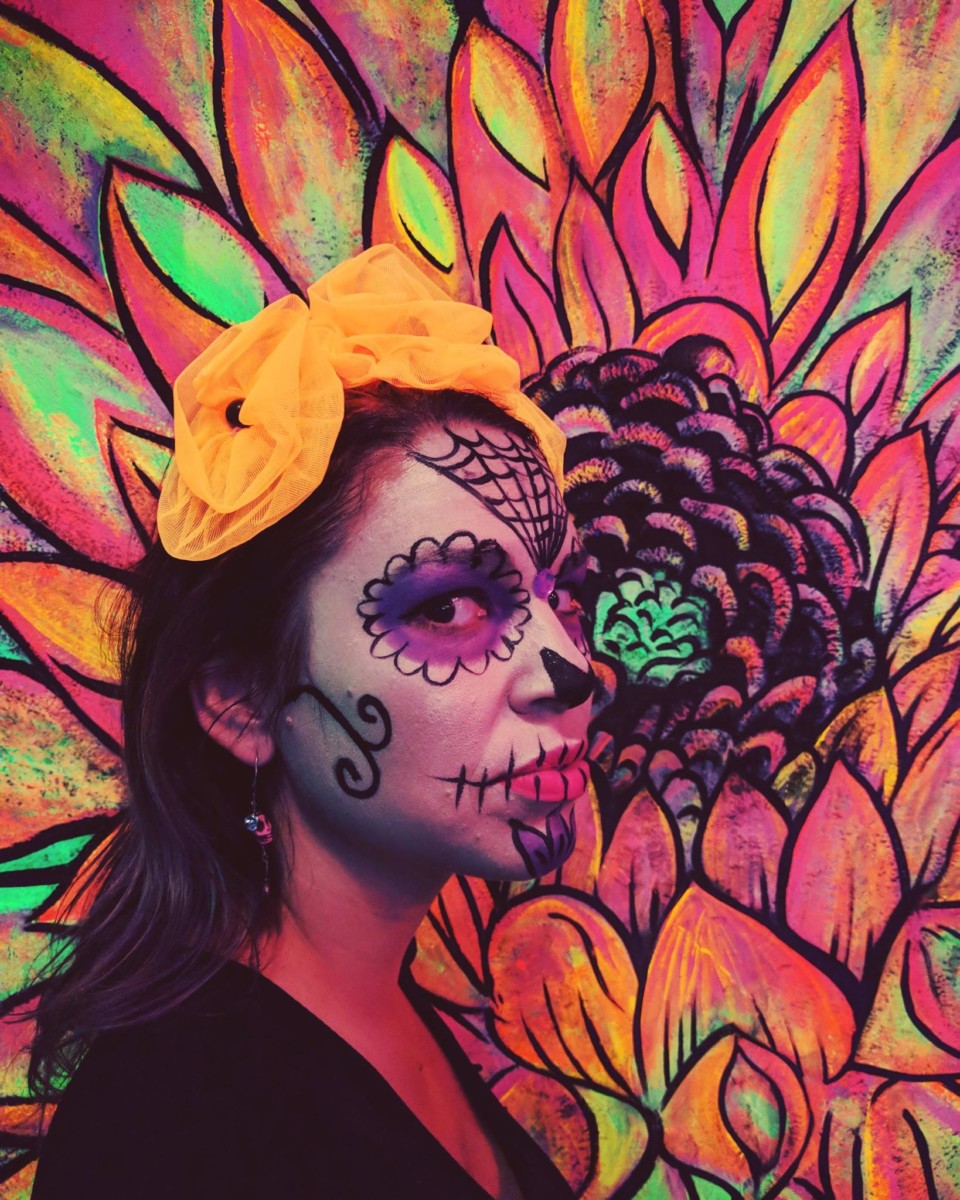 Lolly stands with her face painted in white and black and stands in front of a colorful painting of a flower