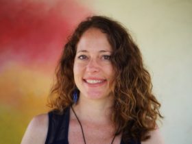 Nora Dunn stands smiling in front of a multi-colored background with reds and yellows