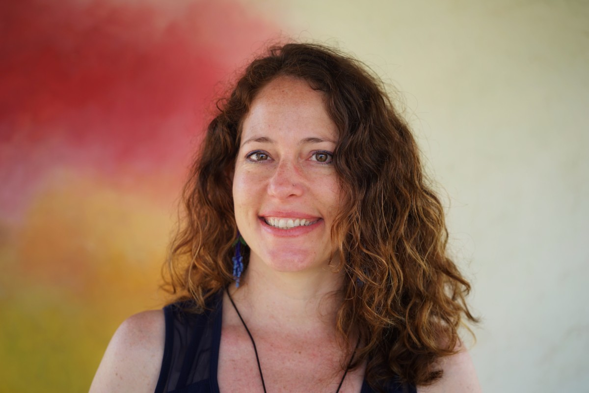 Nora Dunn stands smiling in front of a multi-colored background with reds and yellows