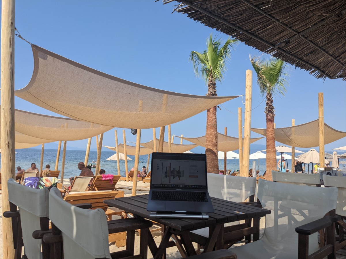 A laptop sits on a table in a restaurant in Greece with palm trees and the ocean nearby